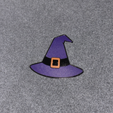 A small witch hat enamel pin in purple with a black band and orange buckle, placed on a textured gray background.