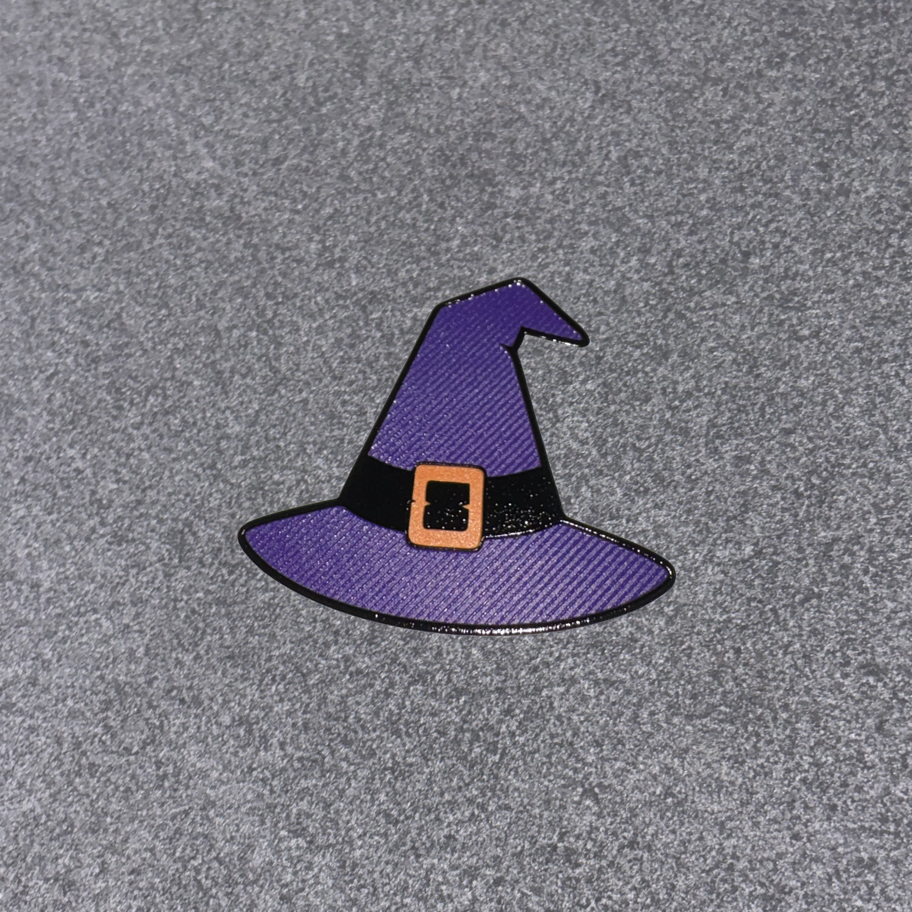 A small witch hat enamel pin in purple with a black band and orange buckle, placed on a textured gray background.