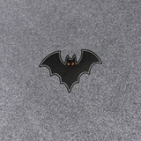 A small bat-shaped patch or emblem with a black body, gray outline, and tiny orange eyes, placed on a textured gray fabric background.