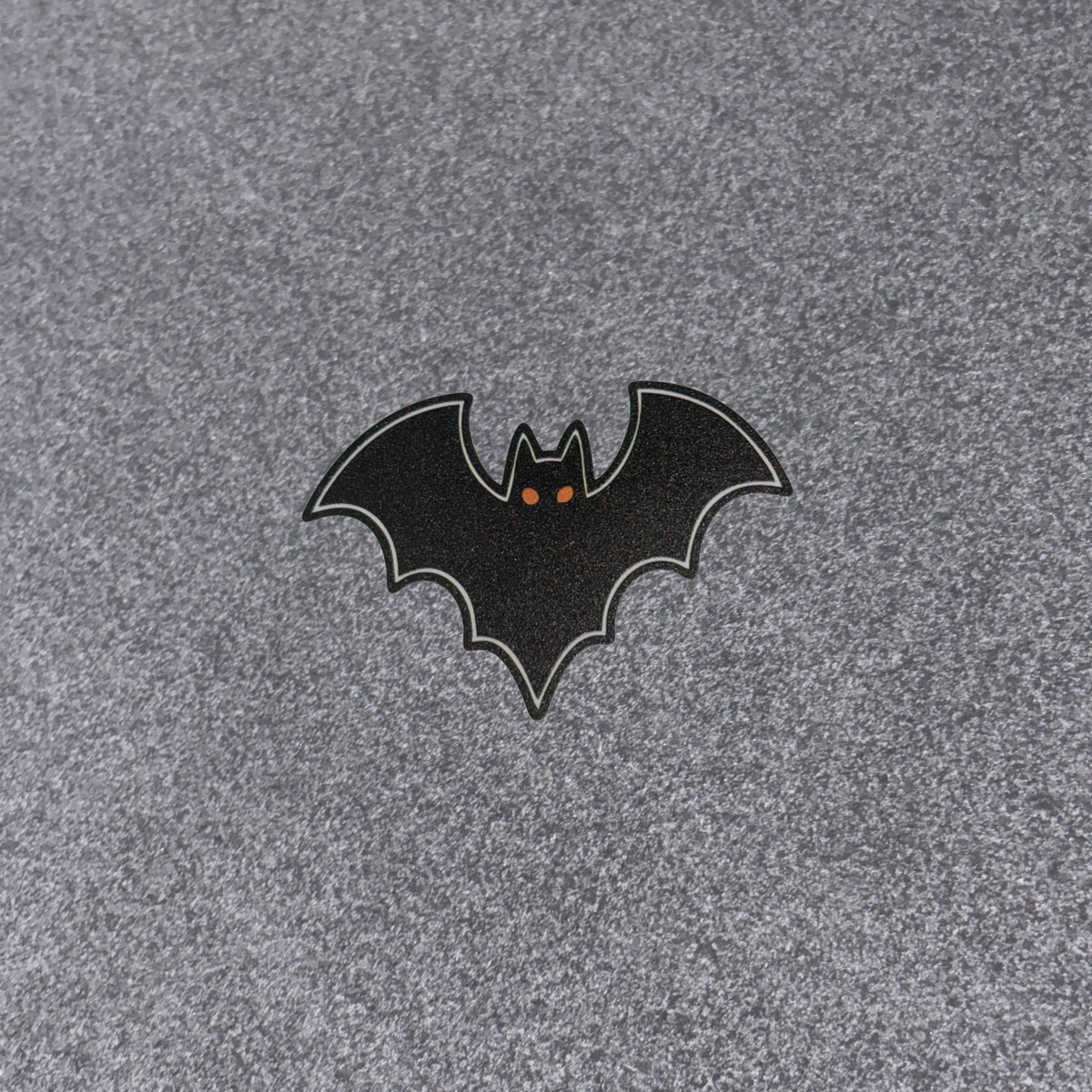 A small bat-shaped patch or emblem with a black body, gray outline, and tiny orange eyes, placed on a textured gray fabric background.