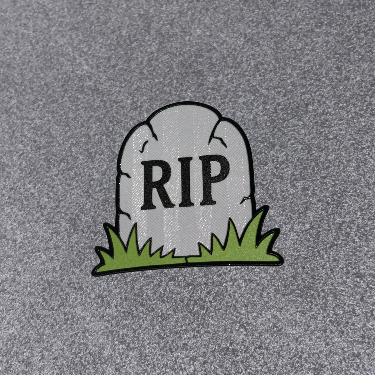 A small tombstone-shaped patch with the letters 'RIP' in black, sitting on green grass, placed on a textured gray background.