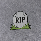 A small tombstone-shaped patch with the letters 'RIP' in black, sitting on green grass, placed on a textured gray background.