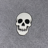 A white skull patch with black details and a black border, placed on a textured gray background.