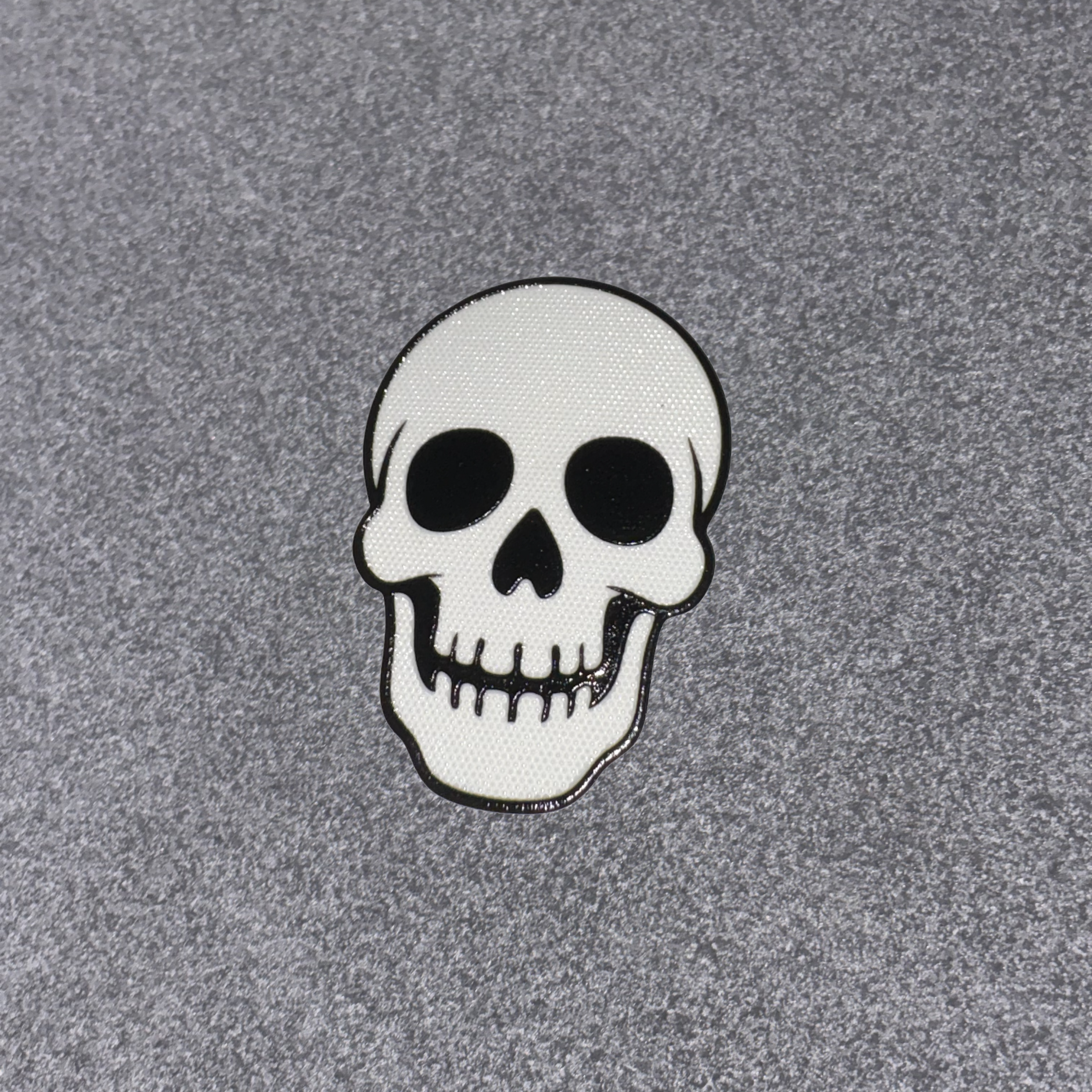 A white skull patch with black details and a black border, placed on a textured gray background.