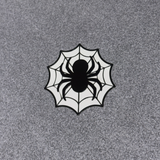 A circular spiderweb patch featuring a black spider silhouette in the center, set against a white web design with black outlines, placed on a textured gray background.