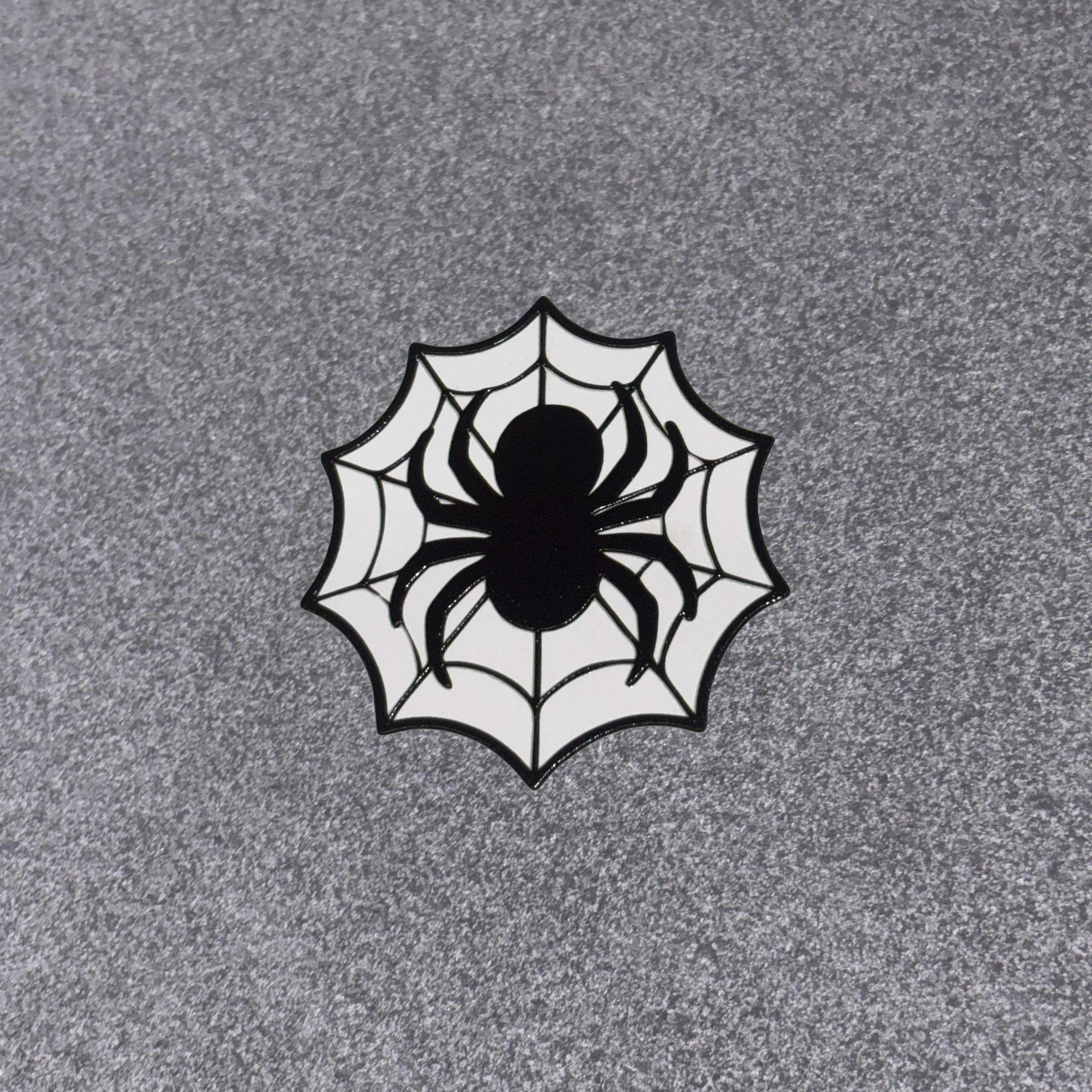 A circular spiderweb patch featuring a black spider silhouette in the center, set against a white web design with black outlines, placed on a textured gray background.
