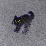 A black cat-shaped accessory with a purple outline and yellow eyes, photographed on a textured gray background.