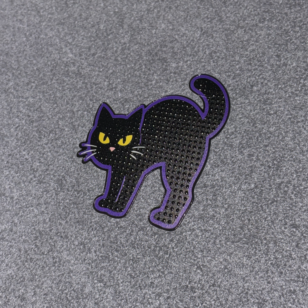 A black cat-shaped accessory with a purple outline and yellow eyes, photographed on a textured gray background.
