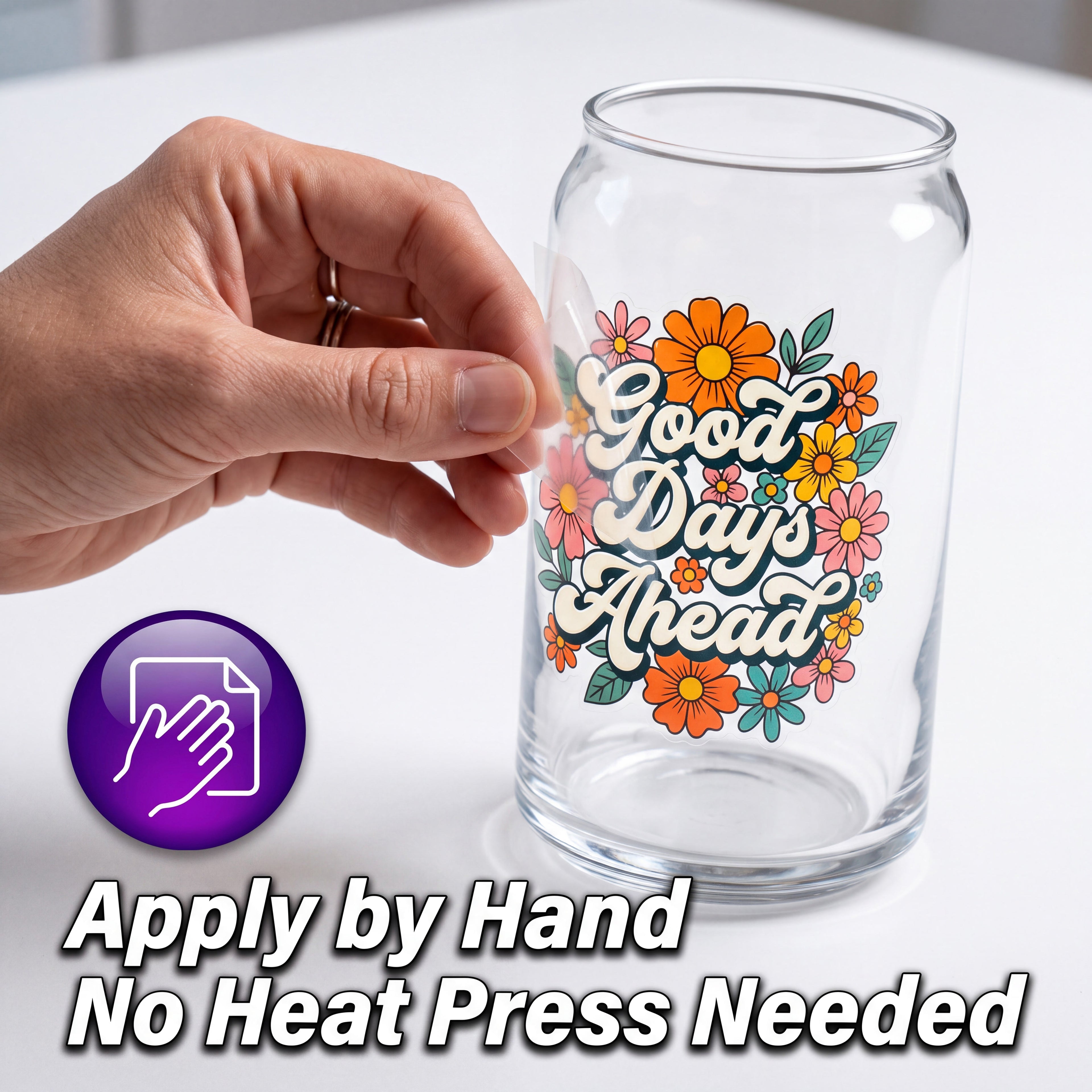 A clear glass can-shaped tumbler with a colorful floral design and the phrase 'Good Days Ahead' in white script, shown on a white surface. A hand is touching the tumbler, and a purple icon plus text at the bottom read 'Apply by Hand' and 'No Heat Press Needed'.