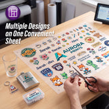 A person's hands hold scissors over a large sheet of assorted printed stickers and decals on a wooden desk. The sheet includes multiple colorful designs such as route-style badges, flowers, animals, and text graphics.